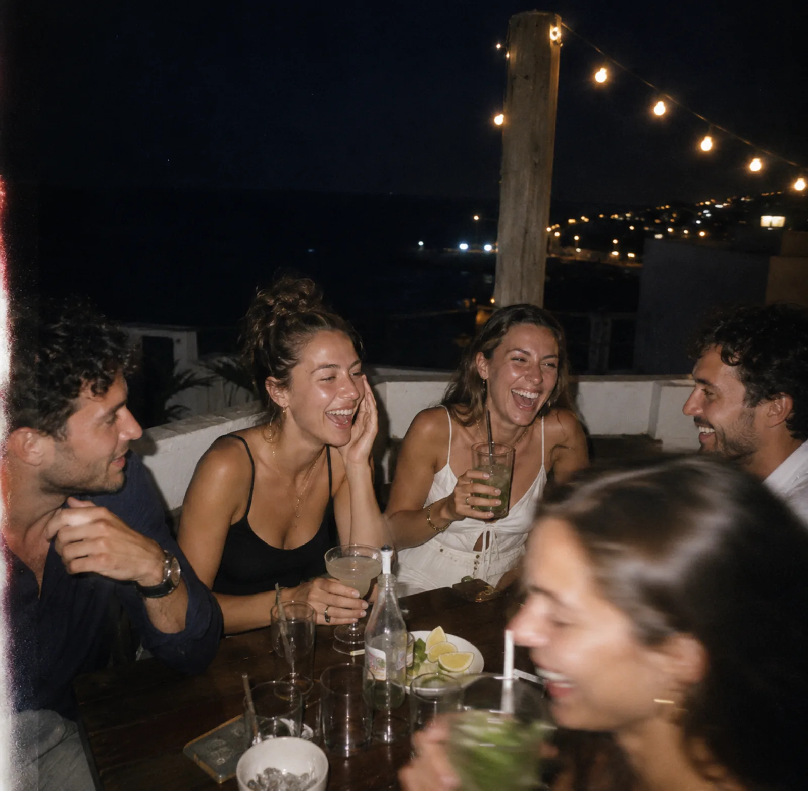 Dîner rooftop nocturne au bord de mer, cinq amis riant autour de cocktails sous une guirlande