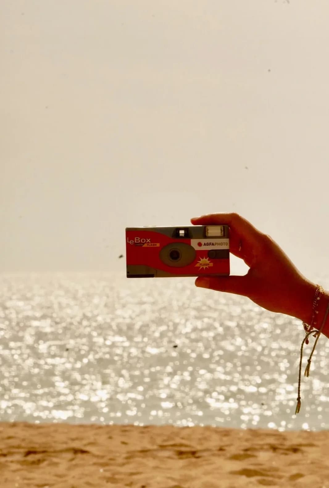 A hand holding a red disposable camera in front of the ocean glittering at sunset