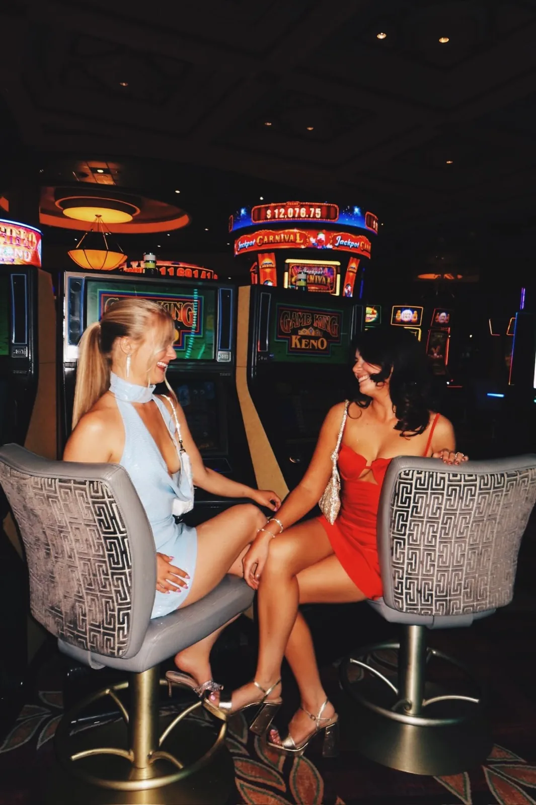Two friends in evening dresses, laughing face to face in front of casino machines
