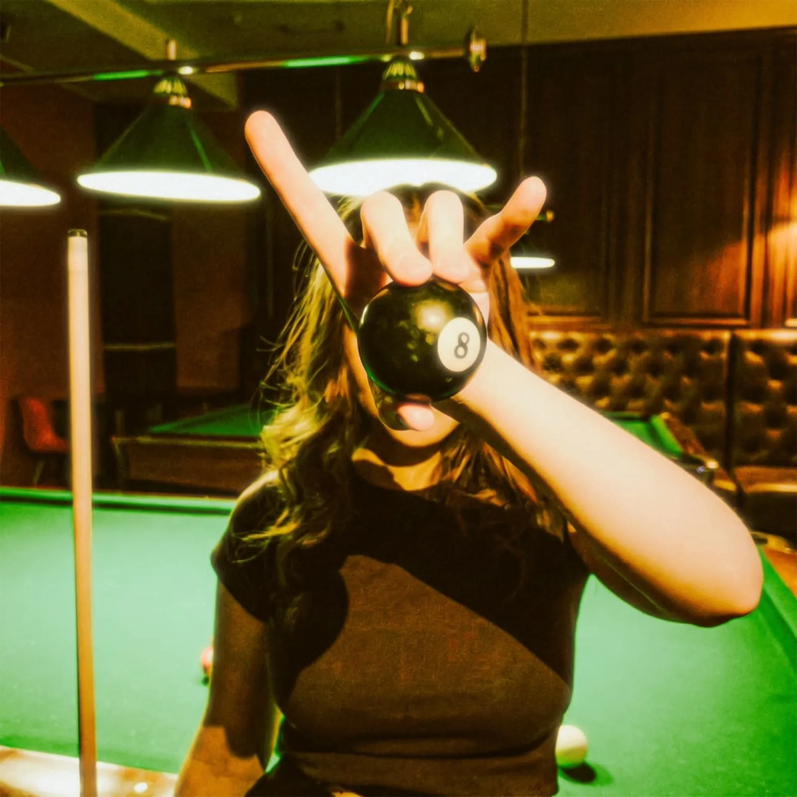 A player holds the black 8-ball up to the lens in a green-lit pool hall