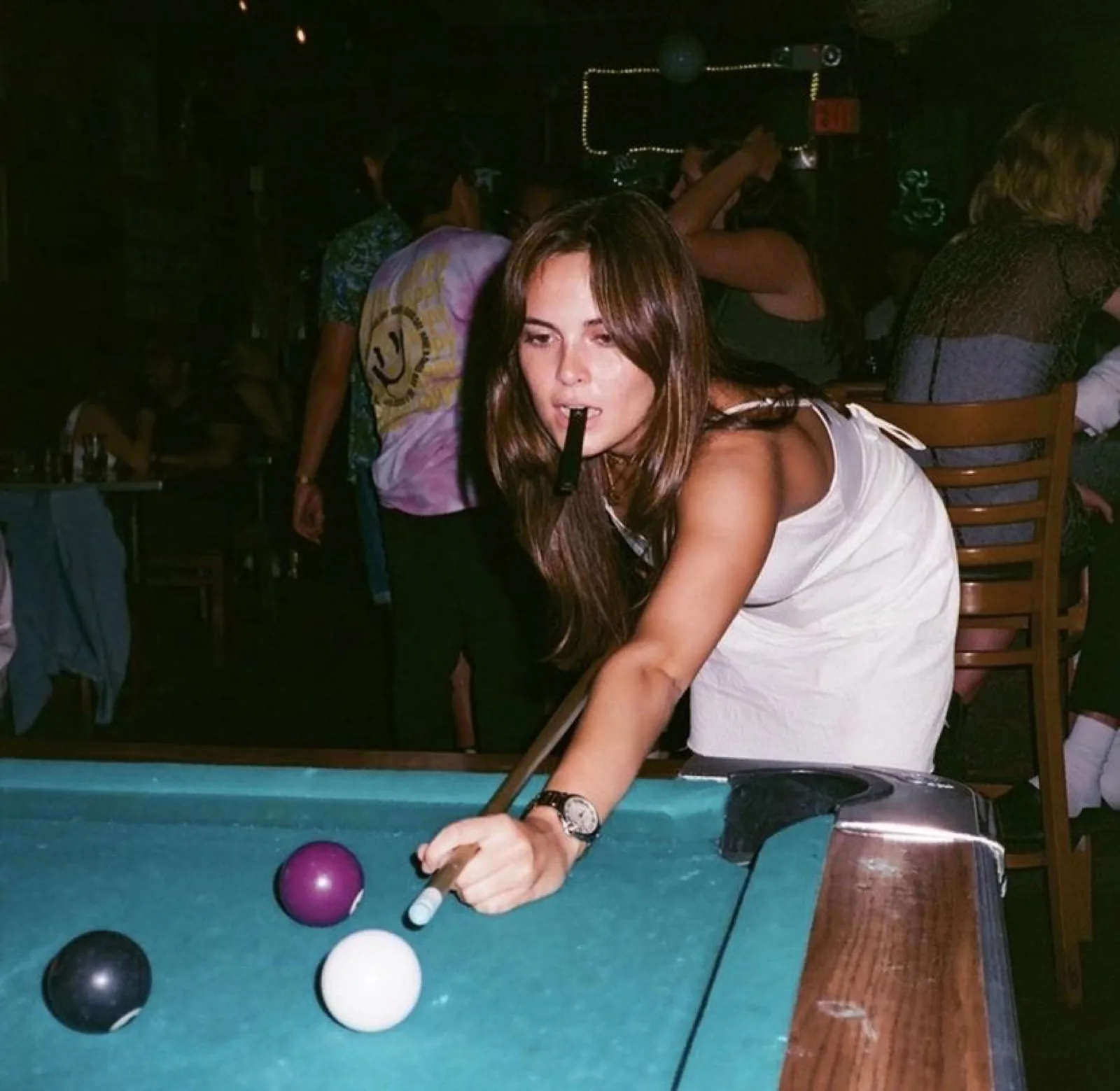A player leaning over a turquoise pool table, cue in hand, a packed bar behind her